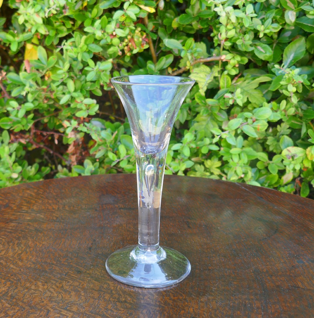 An 18th-century clear hand-blown glass wine goblet with a trumpet bowl, a drawn stem featuring a central teardrop air bubble, and a flat conical base.