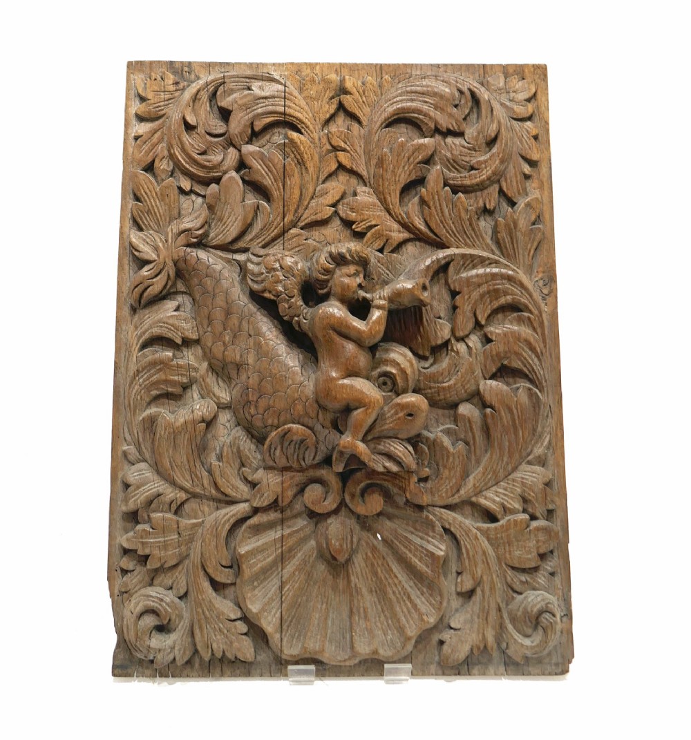 A 17th-century rectangular carved oak relief panel depicting a winged cherub sitting on a dolphin, surrounded by leaf scrolls and a scallop shell. View VI