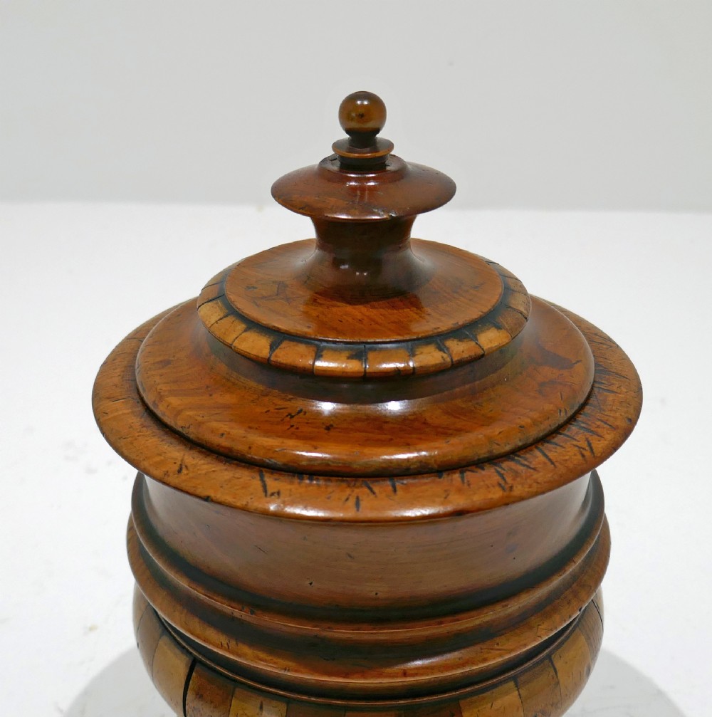A turned antique wooden lidded tobacco jar with staved bands of light and dark timber, set on a pedestal base with small ball feet. View VI