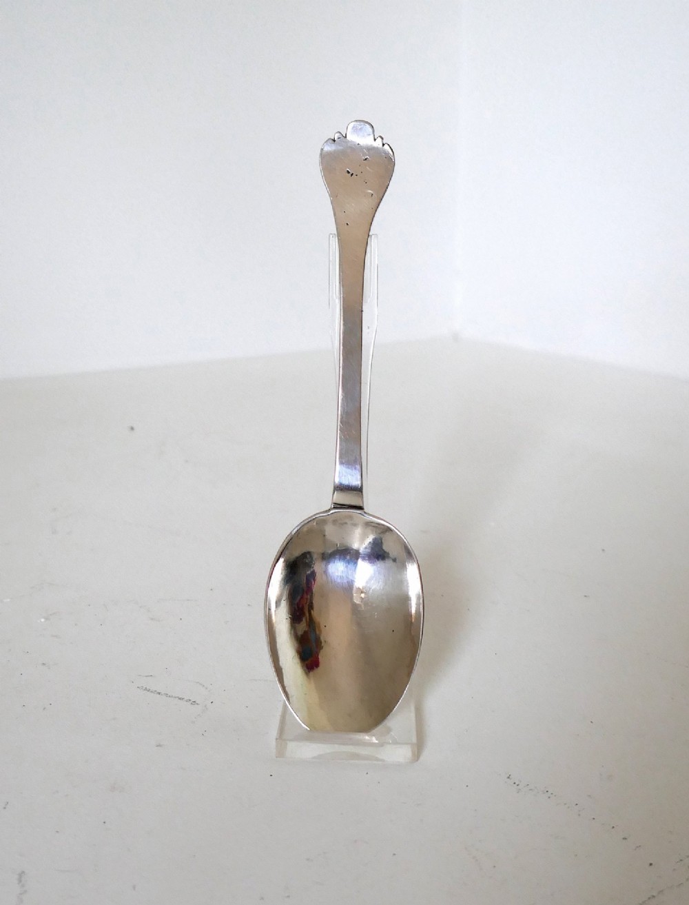 A 17th-century English sterling silver trefid spoon with a lobed terminal handle and an oval bowl displayed on a clear stand.