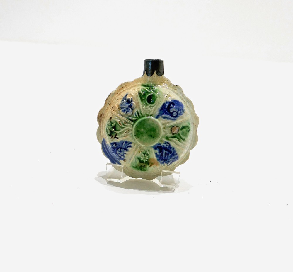 An 18th-century circular ceramic spirit flask featuring relief-moulded floral panels glazed in green, blue, and brown hues on a light cream earthenware body. View II