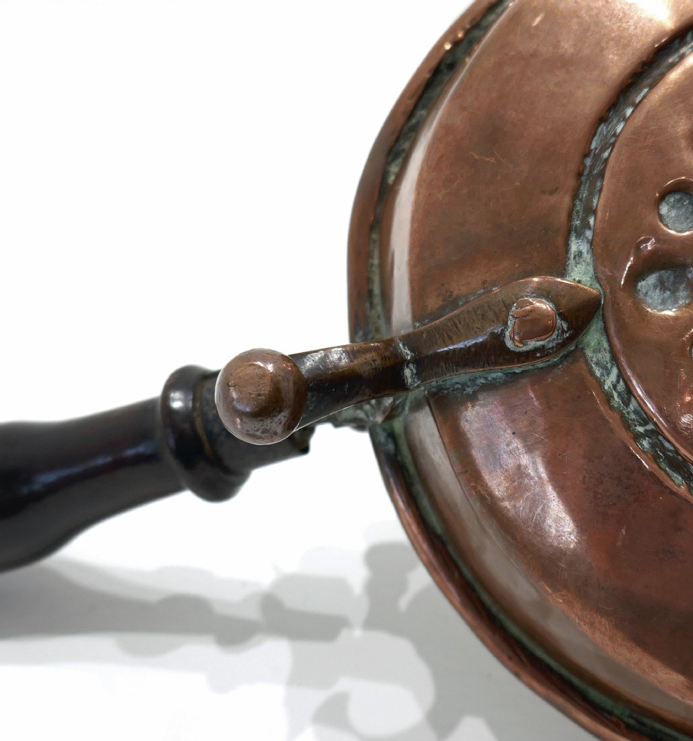 An antique Dutch copper brazier featuring a central repousse motif, a turned fruitwood handle, and three upright rim pegs. View III
