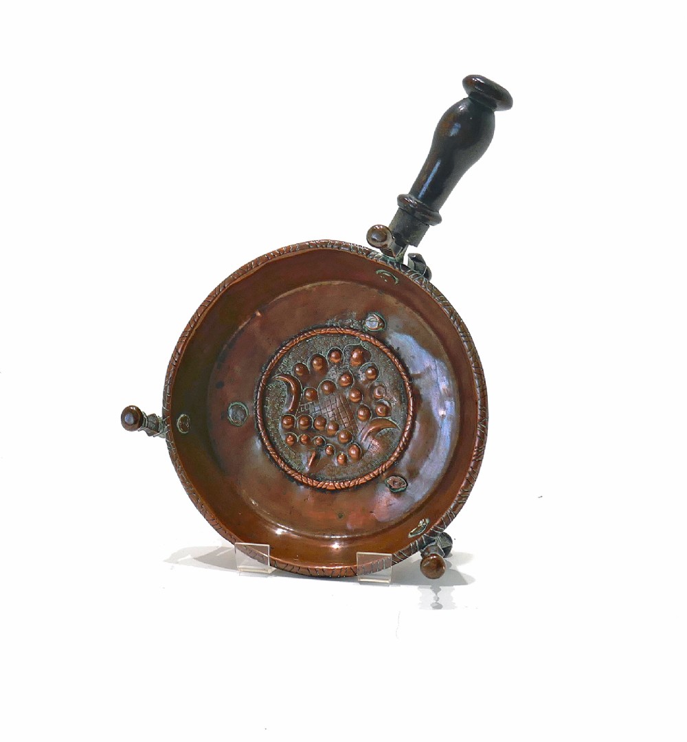 An antique Dutch copper brazier featuring a central repousse motif, a turned fruitwood handle, and three upright rim pegs. View IV