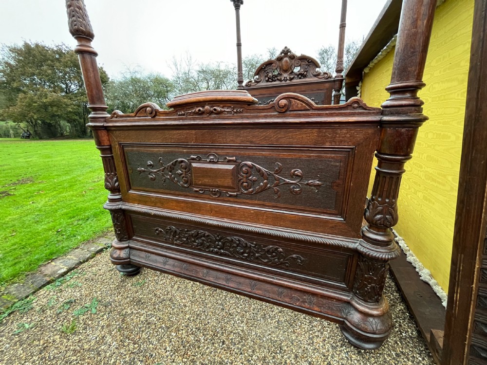 A dark carved oak four poster bed featuring turned columns, an ornate carved headboard, and a gold fabric-lined canopy. View IV