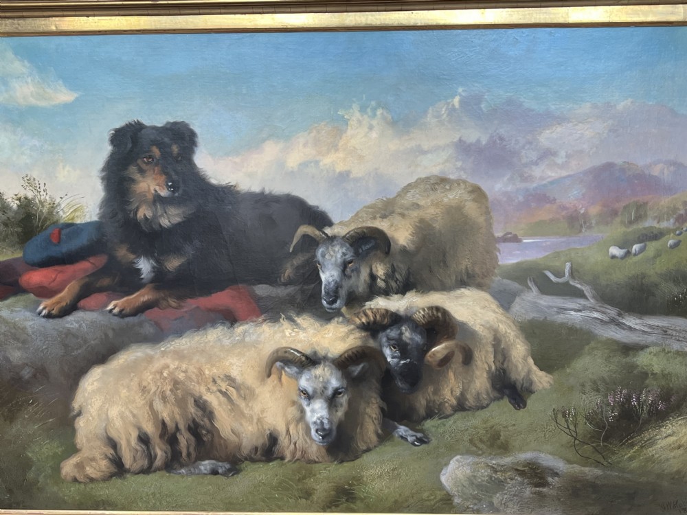 An antique oil painting depicting a black and tan sheepdog resting on a red plaid blanket alongside horned sheep against a mountainous Highland landscape in a gilt frame.