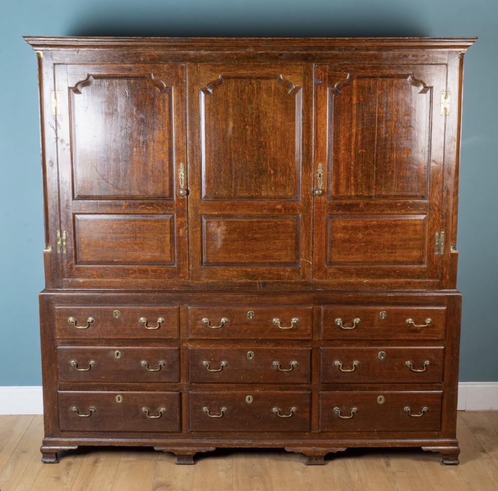A large 18th-century Georgian dark oak press cupboard featuring an upper three-door cabinet with fielded panels over a lower base with nine brass-handled drawers.
