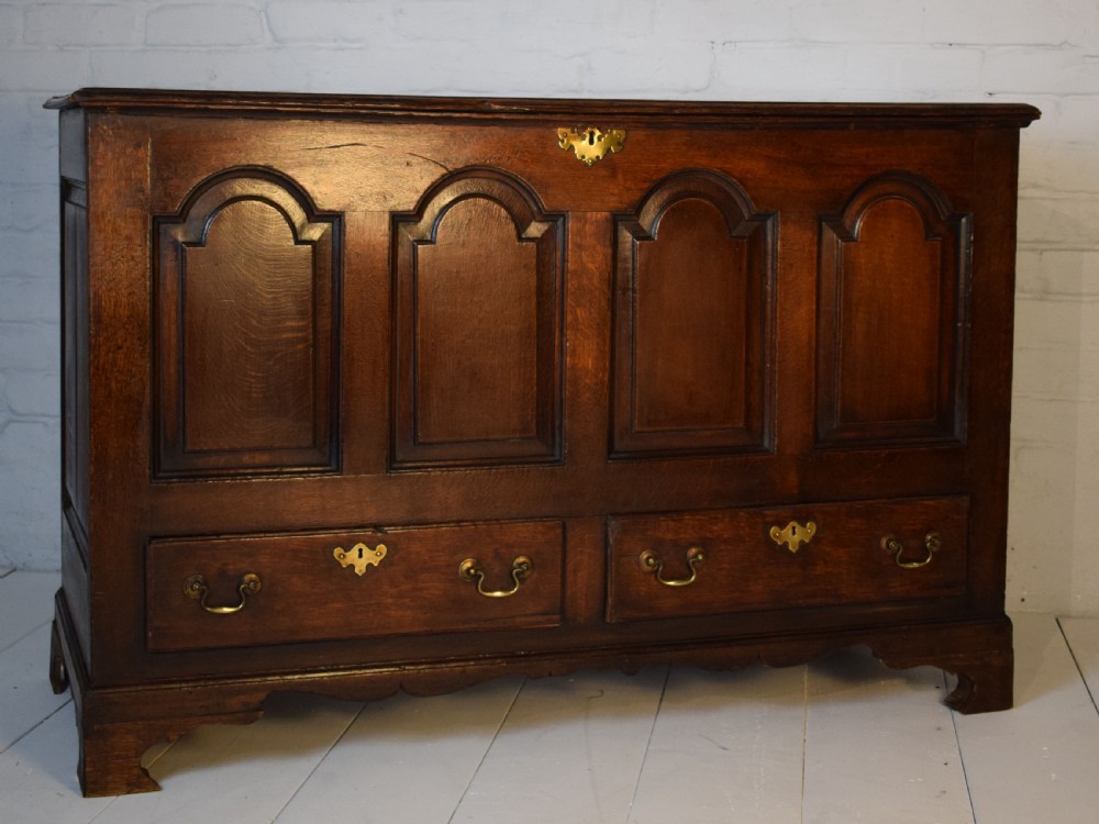 A dark oak antique mule chest featuring four arched fielded panels, two lower drawers with brass drop handles, and bracket feet.