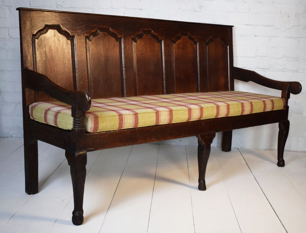 A dark oak Georgian settle featuring a five-panel fielded backrest, shaped armrests, turned legs, and a check pattern seat cushion.