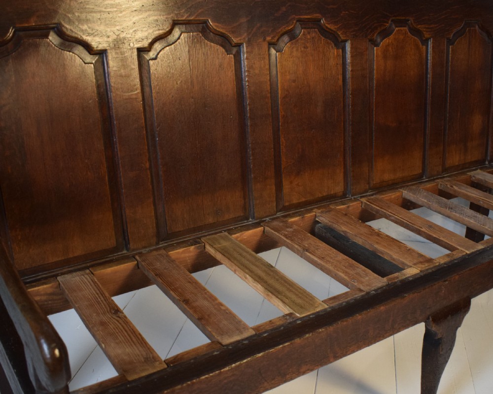A dark oak Georgian settle featuring a five-panel fielded backrest, shaped armrests, turned legs, and a check pattern seat cushion. View IV