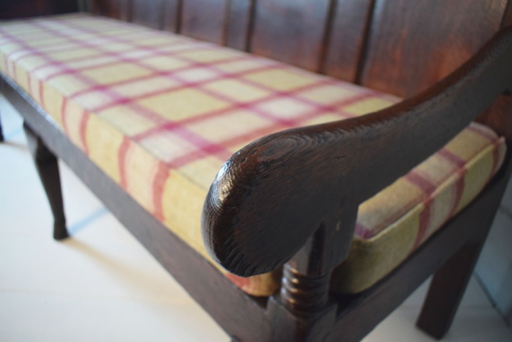 A dark oak Georgian settle featuring a five-panel fielded backrest, shaped armrests, turned legs, and a check pattern seat cushion. View VII
