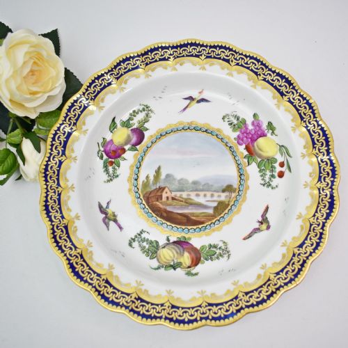 fig 1. An antique circular porcelain plate featuring a scalloped gilded rim, cobalt blue border, hand-painted fruit and bird motifs, and a central landscape roundel.