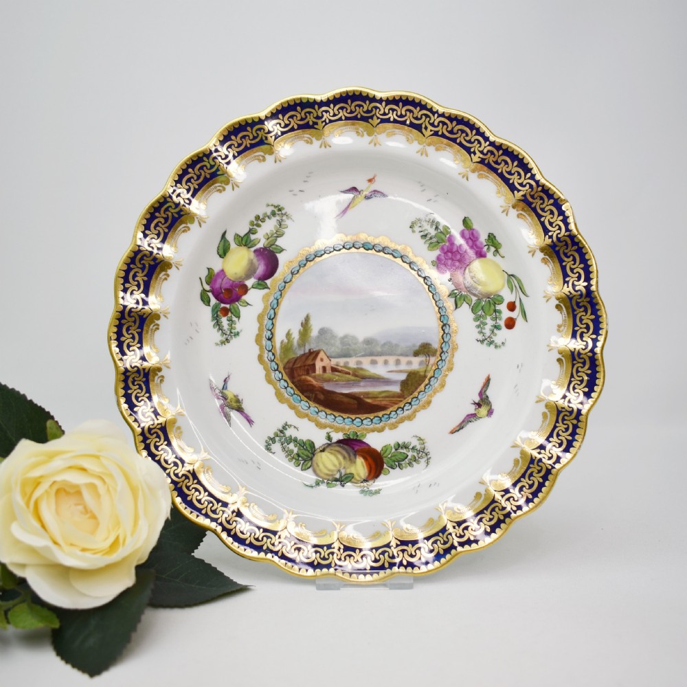 An antique circular porcelain plate featuring a scalloped gilded rim, cobalt blue border, hand-painted fruit and bird motifs, and a central landscape roundel.