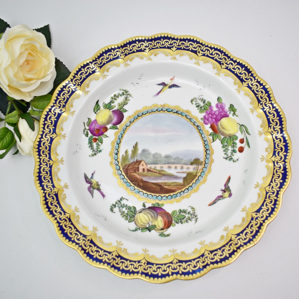 An antique circular porcelain plate featuring a scalloped gilded rim, cobalt blue border, hand-painted fruit and bird motifs, and a central landscape roundel. View II