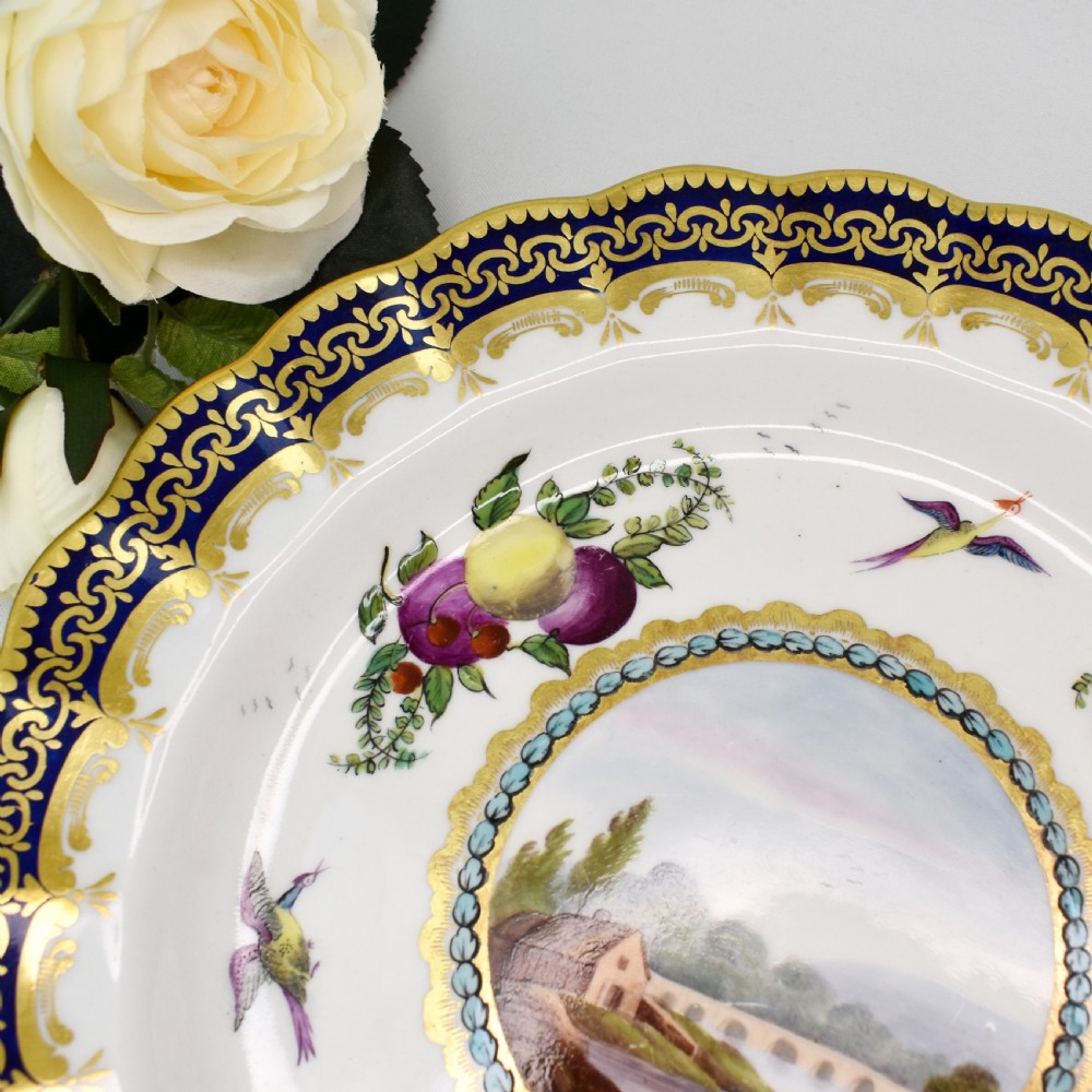 An antique circular porcelain plate featuring a scalloped gilded rim, cobalt blue border, hand-painted fruit and bird motifs, and a central landscape roundel. View III
