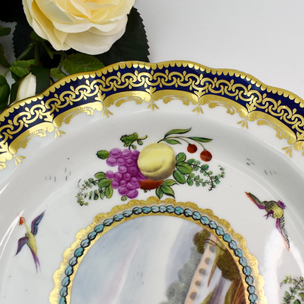 An antique circular porcelain plate featuring a scalloped gilded rim, cobalt blue border, hand-painted fruit and bird motifs, and a central landscape roundel. View IV