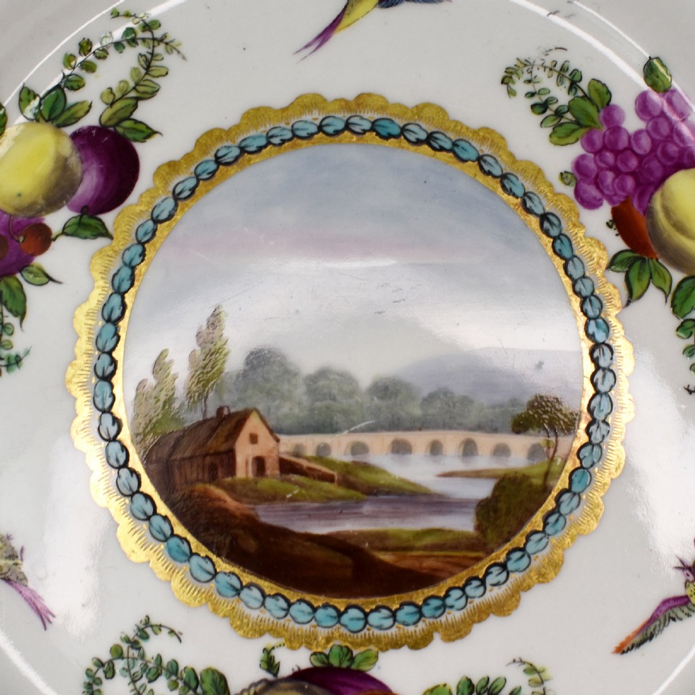 An antique circular porcelain plate featuring a scalloped gilded rim, cobalt blue border, hand-painted fruit and bird motifs, and a central landscape roundel. View V
