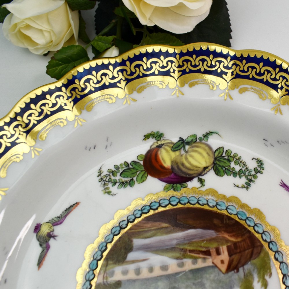 An antique circular porcelain plate featuring a scalloped gilded rim, cobalt blue border, hand-painted fruit and bird motifs, and a central landscape roundel. View VI