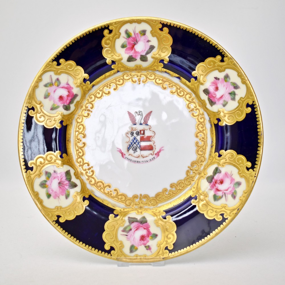 A circular porcelain dessert plate featuring a cobalt blue rim with six gilt-framed reserves depicting hand-painted pink roses around a central family coat of arms.