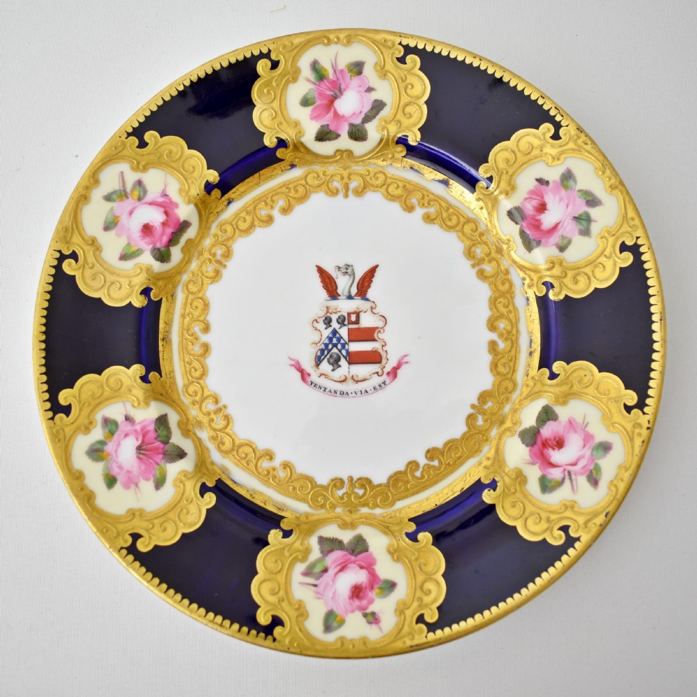 A circular porcelain dessert plate featuring a cobalt blue rim with six gilt-framed reserves depicting hand-painted pink roses around a central family coat of arms. View II
