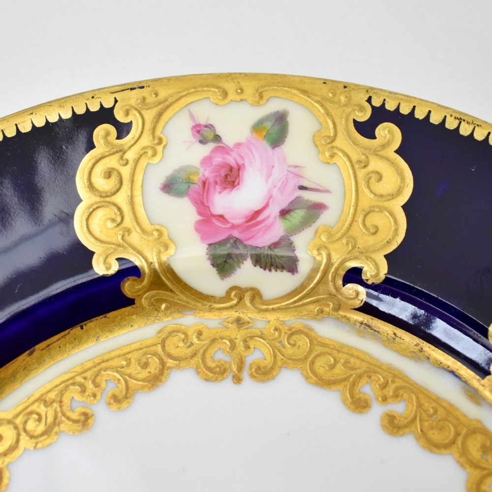 A circular porcelain dessert plate featuring a cobalt blue rim with six gilt-framed reserves depicting hand-painted pink roses around a central family coat of arms. View III