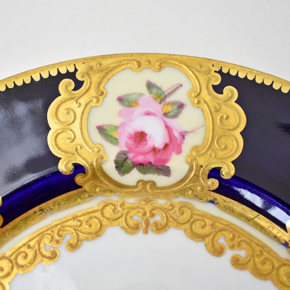 A circular porcelain dessert plate featuring a cobalt blue rim with six gilt-framed reserves depicting hand-painted pink roses around a central family coat of arms. View VI
