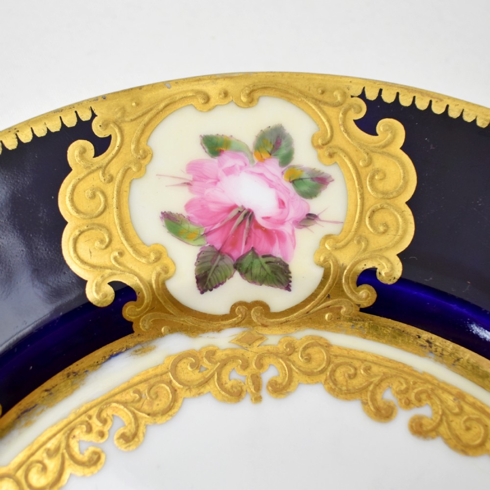 A circular porcelain dessert plate featuring a cobalt blue rim with six gilt-framed reserves depicting hand-painted pink roses around a central family coat of arms. View VII