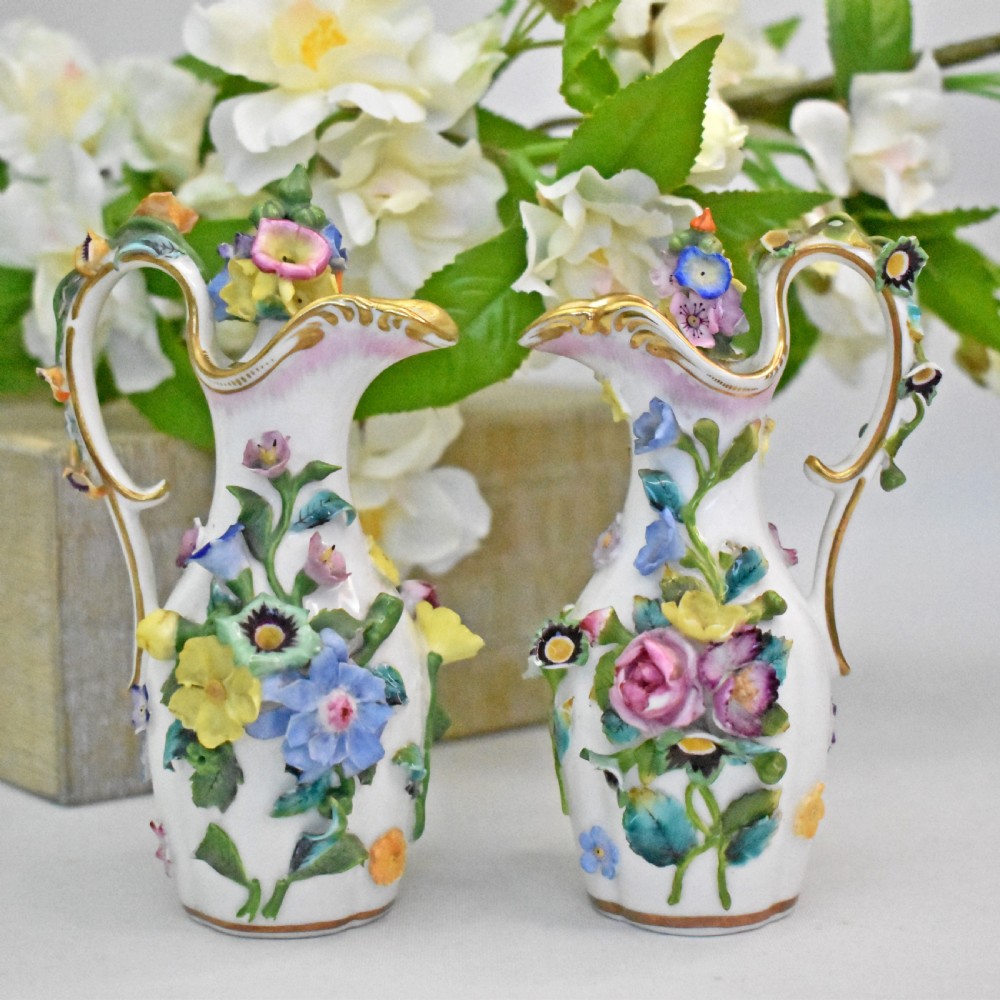 A pair of white Minton porcelain scent bottle ewers featuring applied three-dimensional flowers, gilded scroll handles, and matching floral stoppers.