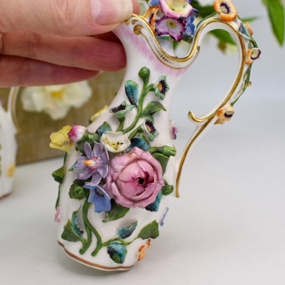 A pair of white Minton porcelain scent bottle ewers featuring applied three-dimensional flowers, gilded scroll handles, and matching floral stoppers. View V