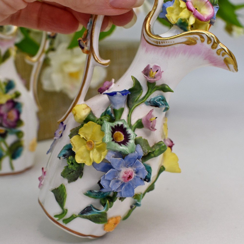 A pair of white Minton porcelain scent bottle ewers featuring applied three-dimensional flowers, gilded scroll handles, and matching floral stoppers. View VI