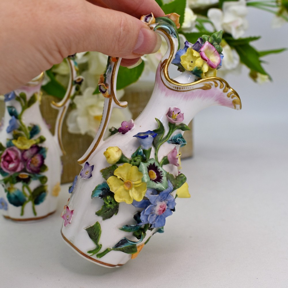 A pair of white Minton porcelain scent bottle ewers featuring applied three-dimensional flowers, gilded scroll handles, and matching floral stoppers. View VIII