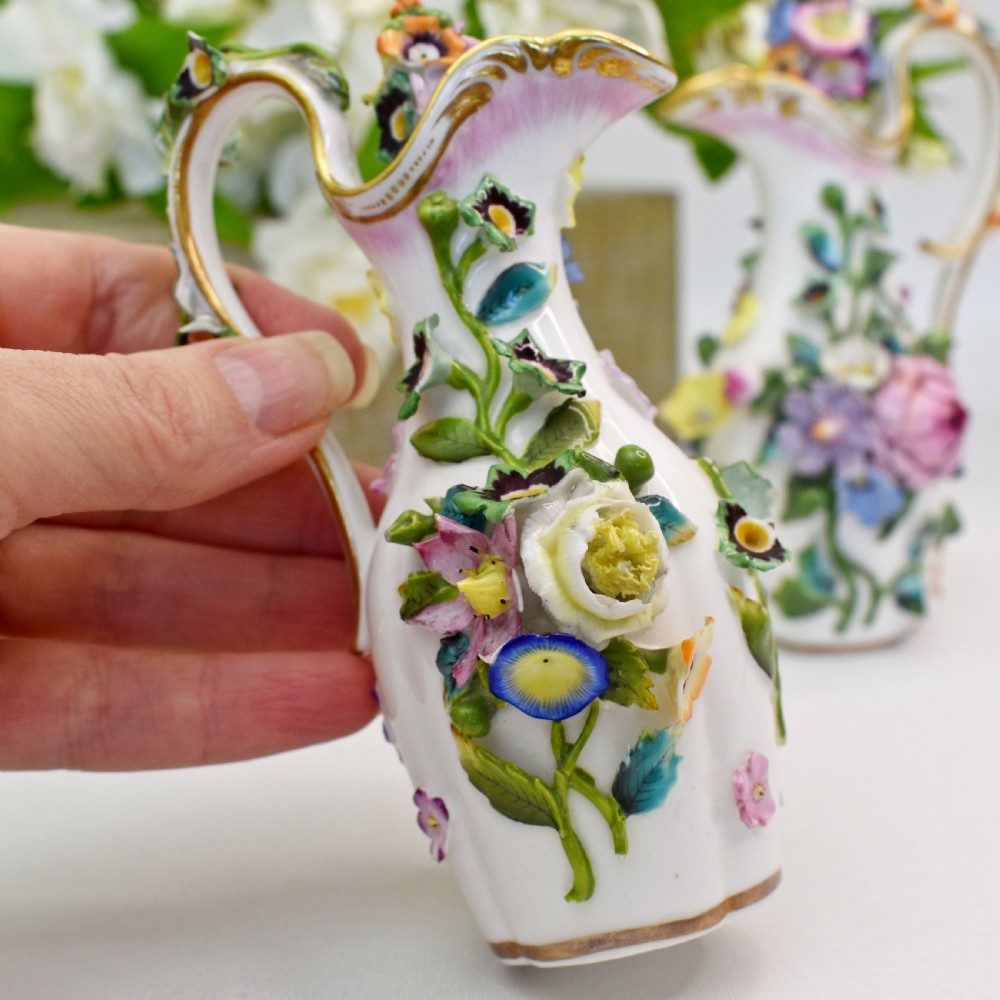 A pair of white Minton porcelain scent bottle ewers featuring applied three-dimensional flowers, gilded scroll handles, and matching floral stoppers. View IX