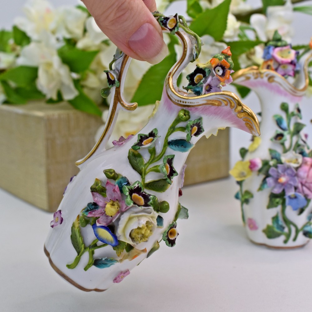 A pair of white Minton porcelain scent bottle ewers featuring applied three-dimensional flowers, gilded scroll handles, and matching floral stoppers. View X