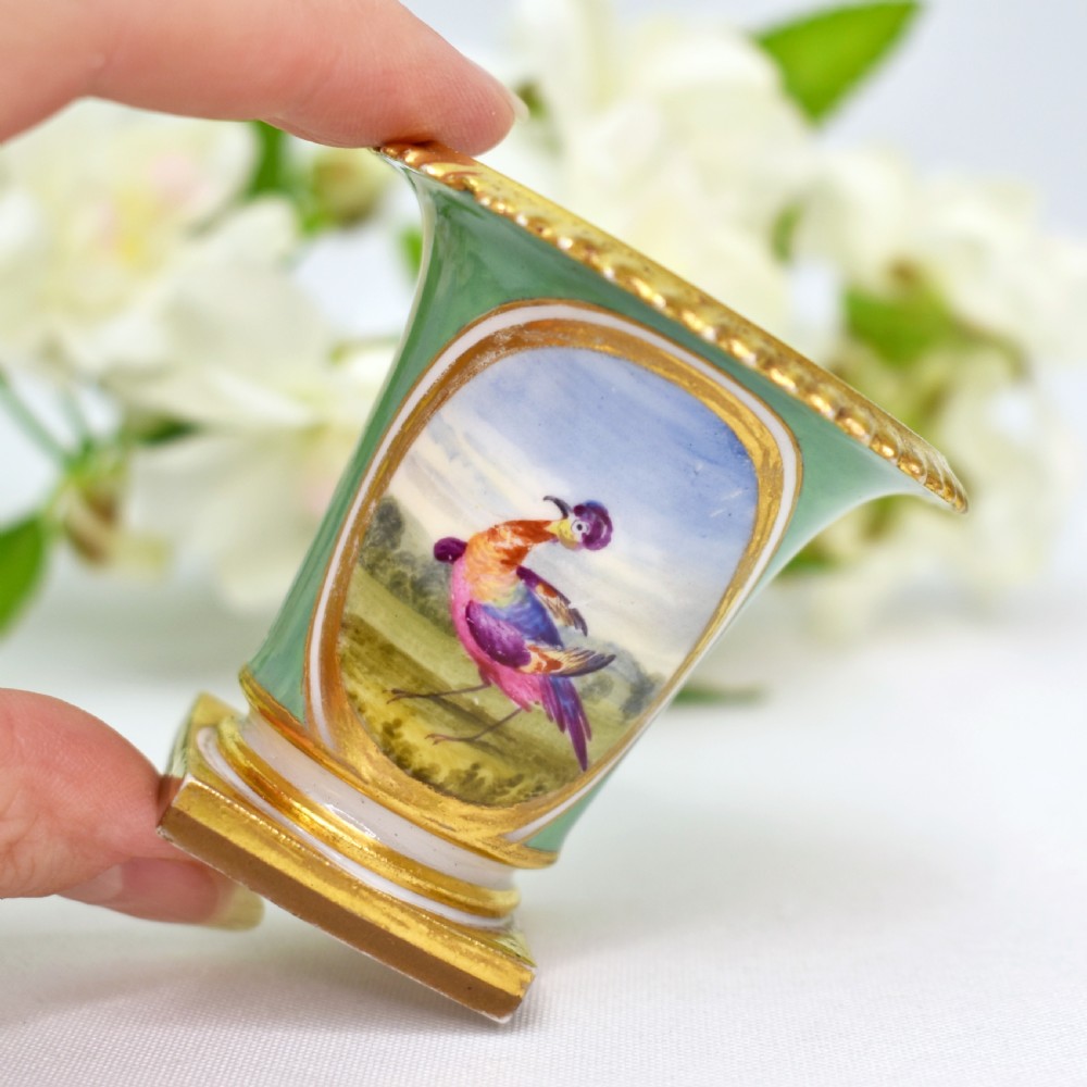 A miniature green Regency porcelain vase featuring a hand-painted exotic bird vignette framed in gold gilding upon a square pedestal base. View VI