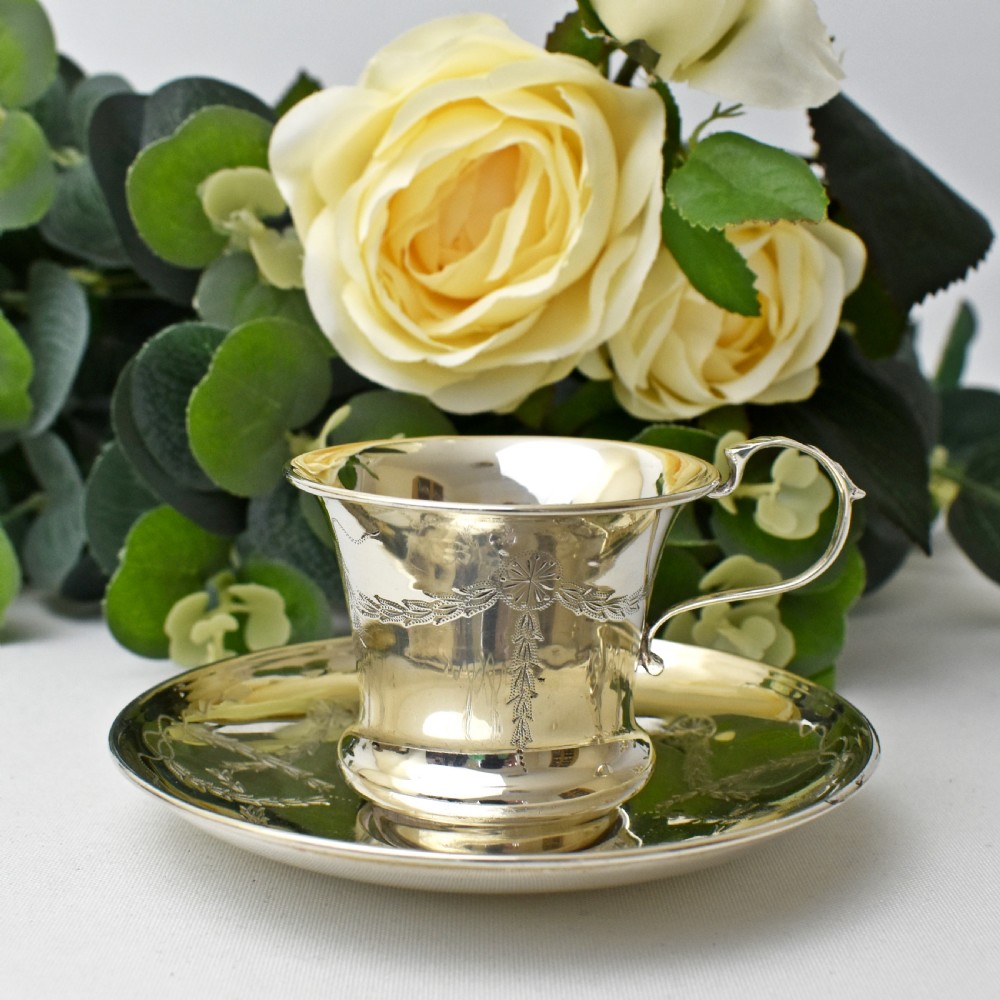 An antique sterling silver cup and saucer set decorated with hand-engraved swag and bow motifs.