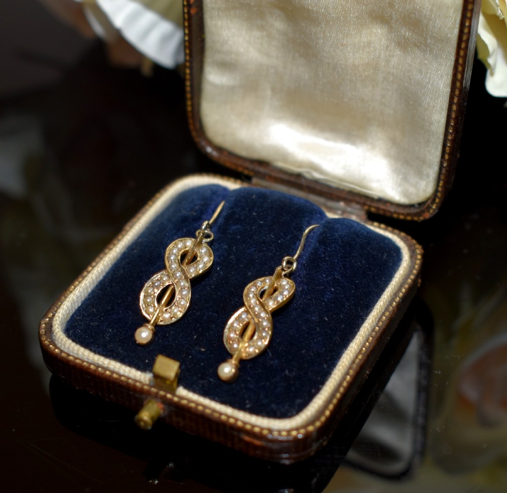 A pair of Edwardian gold drop earrings featuring figure-eight infinity loops set with seed pearls, displayed inside a blue velvet presentation box.