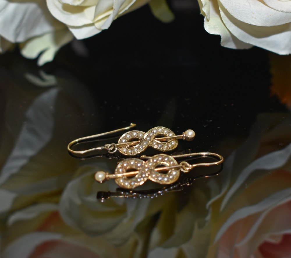 A pair of Edwardian gold drop earrings featuring figure-eight infinity loops set with seed pearls, displayed inside a blue velvet presentation box. View III