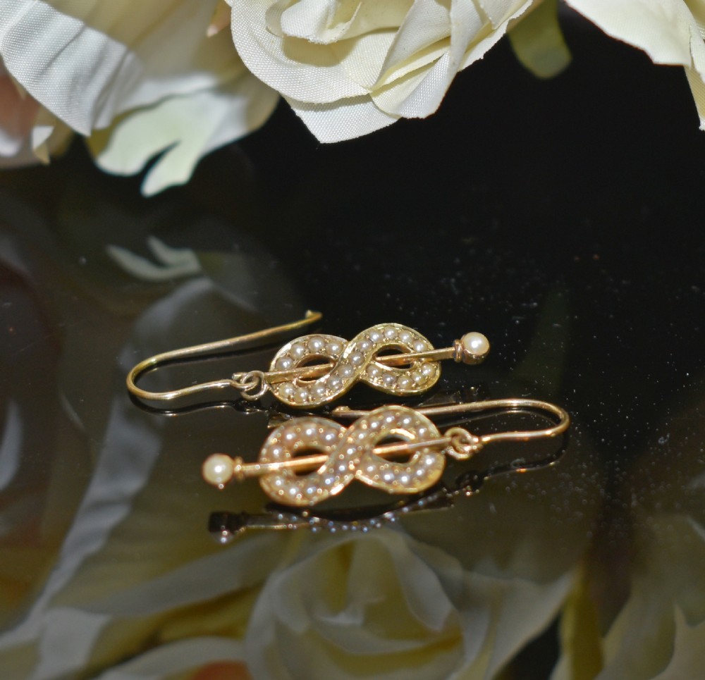 A pair of Edwardian gold drop earrings featuring figure-eight infinity loops set with seed pearls, displayed inside a blue velvet presentation box. View V