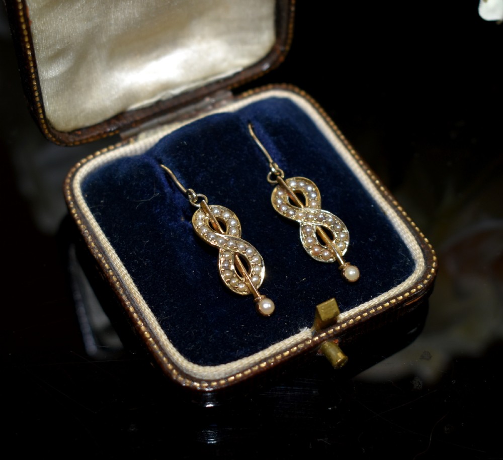 A pair of Edwardian gold drop earrings featuring figure-eight infinity loops set with seed pearls, displayed inside a blue velvet presentation box. View VI