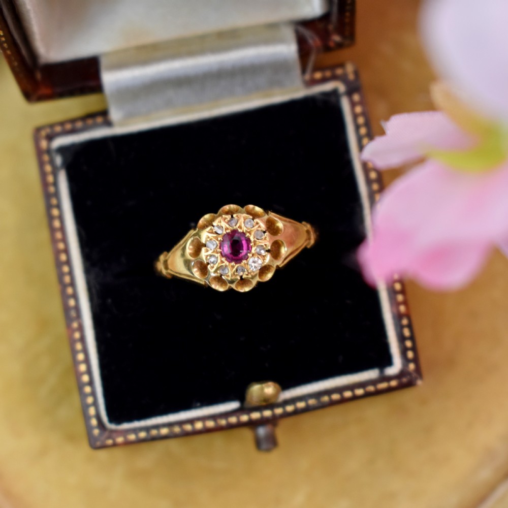 An 18ct yellow gold Edwardian ring featuring a central round ruby encircled by diamond accents set into a carved head band.