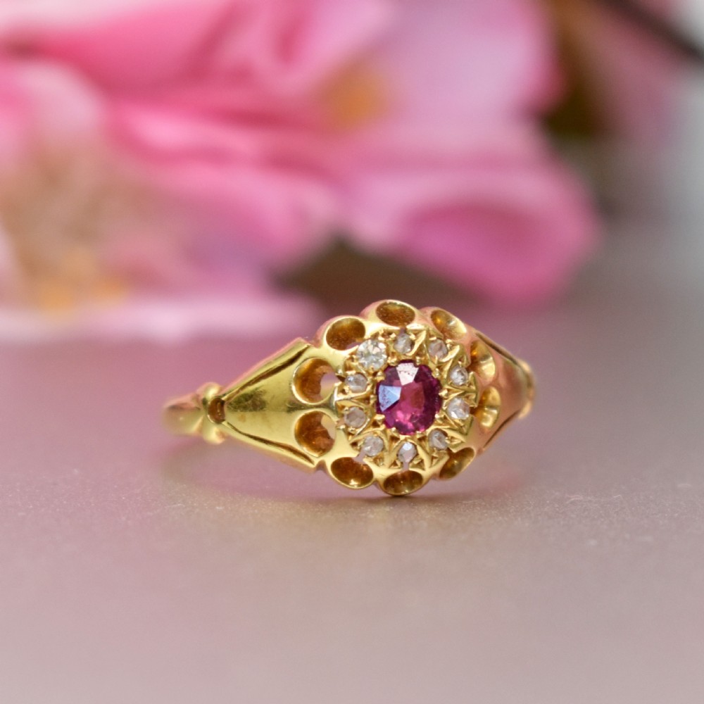 An 18ct yellow gold Edwardian ring featuring a central round ruby encircled by diamond accents set into a carved head band. View V