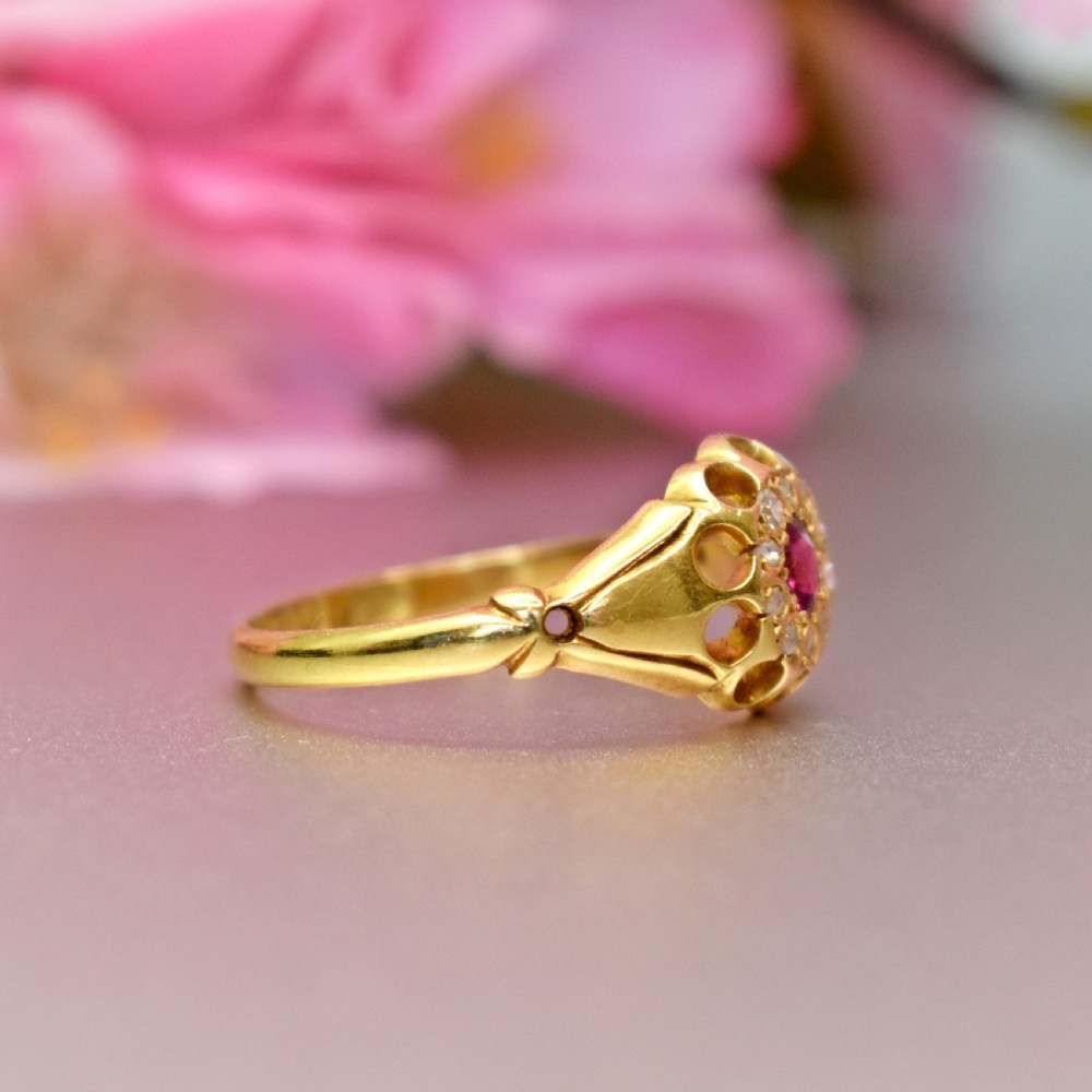 An 18ct yellow gold Edwardian ring featuring a central round ruby encircled by diamond accents set into a carved head band. View VI