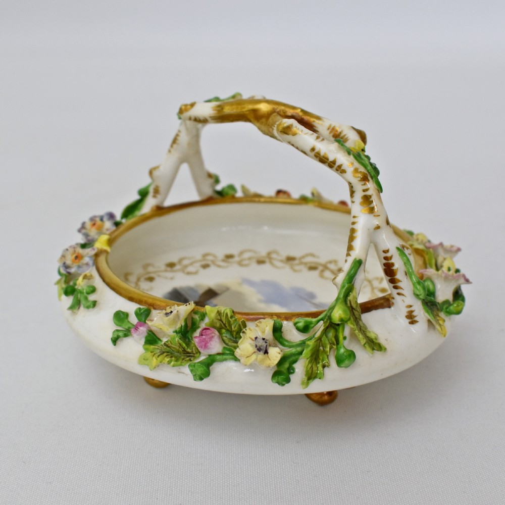 A circular Rockingham porcelain basket featuring applied botanical flowers, a branch-formed gilded handle, and a hand-painted landscape scene at the centre.