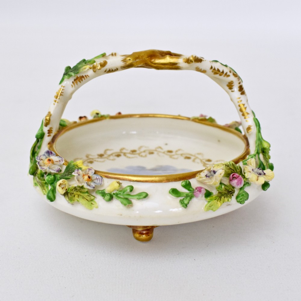 A circular Rockingham porcelain basket featuring applied botanical flowers, a branch-formed gilded handle, and a hand-painted landscape scene at the centre. View II