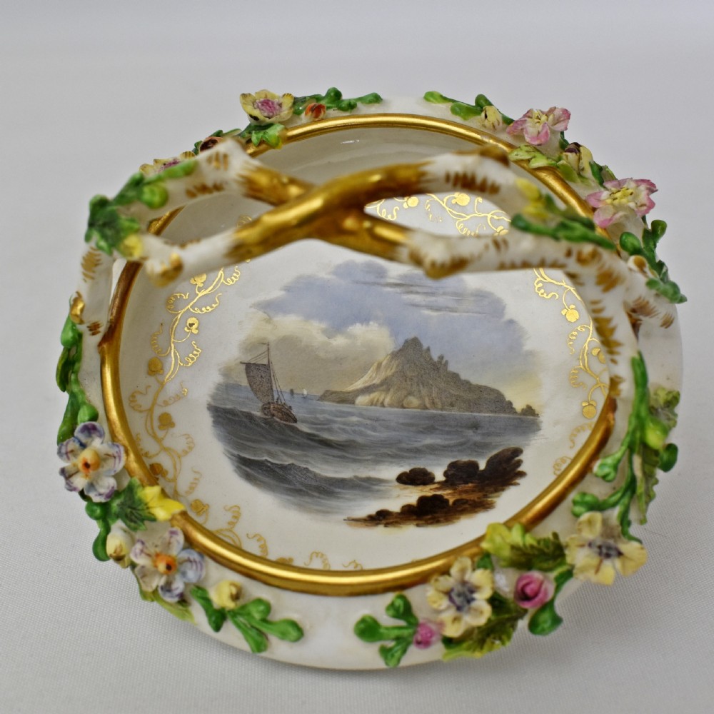 A circular Rockingham porcelain basket featuring applied botanical flowers, a branch-formed gilded handle, and a hand-painted landscape scene at the centre. View III