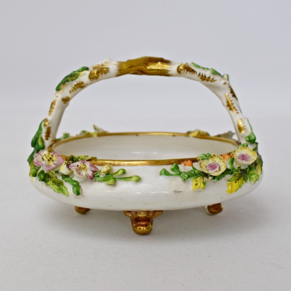 A circular Rockingham porcelain basket featuring applied botanical flowers, a branch-formed gilded handle, and a hand-painted landscape scene at the centre. View VI