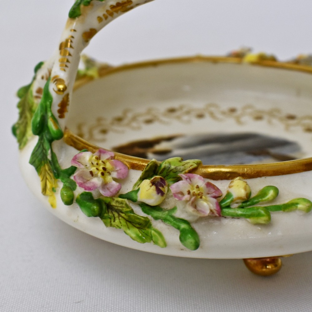 A circular Rockingham porcelain basket featuring applied botanical flowers, a branch-formed gilded handle, and a hand-painted landscape scene at the centre. View VII