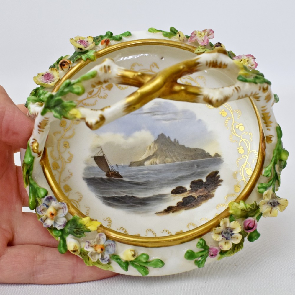 A circular Rockingham porcelain basket featuring applied botanical flowers, a branch-formed gilded handle, and a hand-painted landscape scene at the centre. View VIII