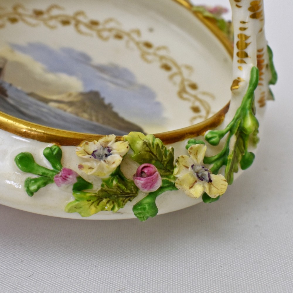A circular Rockingham porcelain basket featuring applied botanical flowers, a branch-formed gilded handle, and a hand-painted landscape scene at the centre. View XI