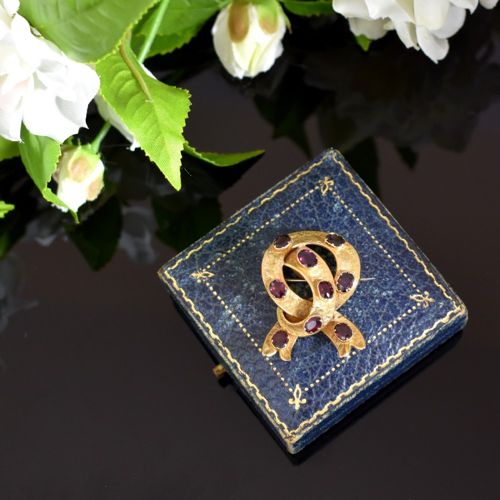 A Victorian 15ct gold brooch styled as an intertwined ribbon loop, textured with hand engraving and set with oval flat-cut almandine garnets. View VI