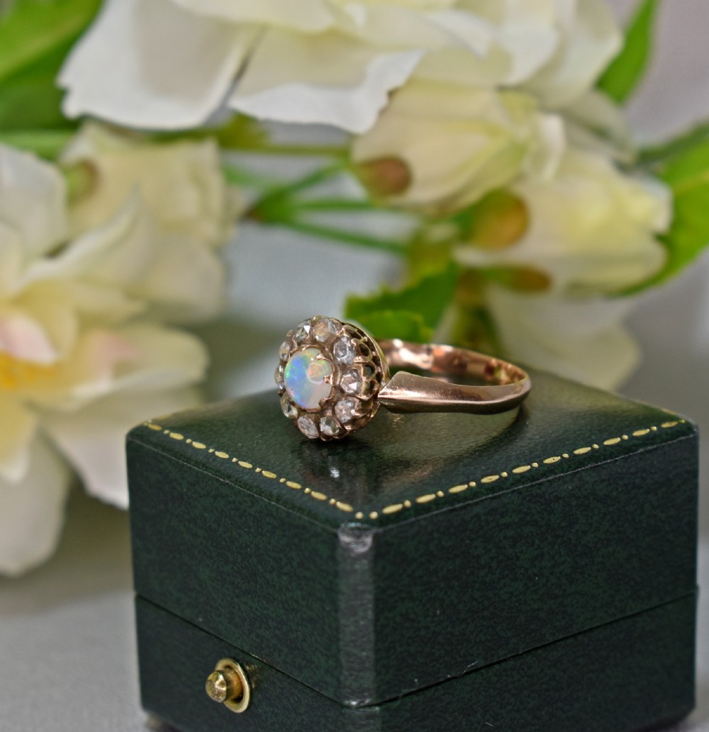 An antique gold Victorian cluster ring featuring a central cabochon opal surrounded by a halo of rose-cut diamonds, displayed inside a green velvet ring box. View V